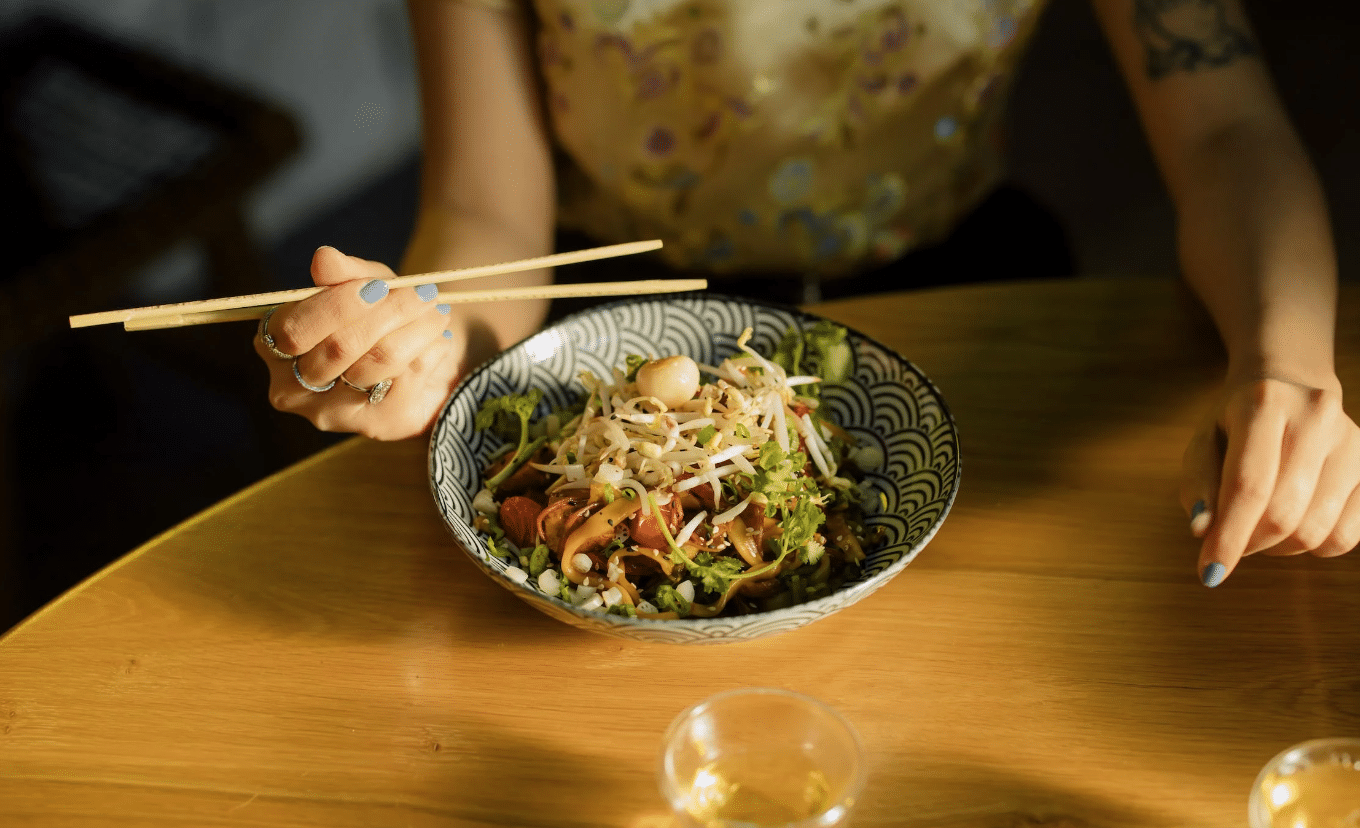 lady with arm tattoo holding chopsticks over a bowl of fresh noodles