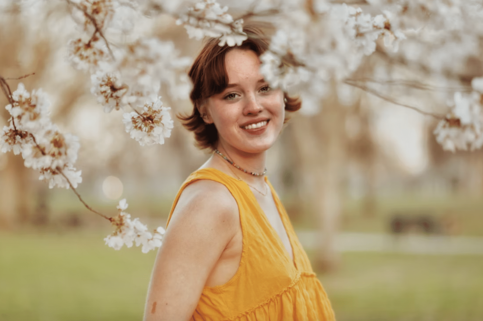 young woman by a blossom tree smiling wearing a yellow sleeveless top