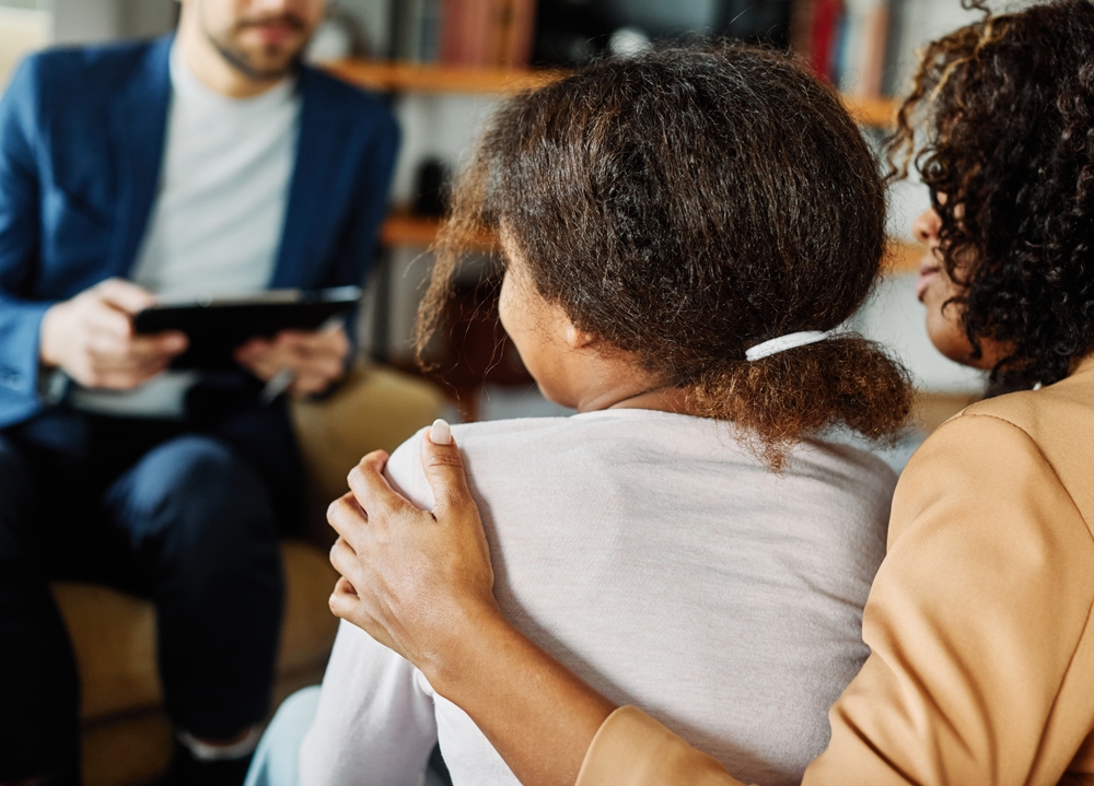 A mother has her arm round her teenage daughter across from a male therapist, depicting that eating disorders in teens affect the whole family.