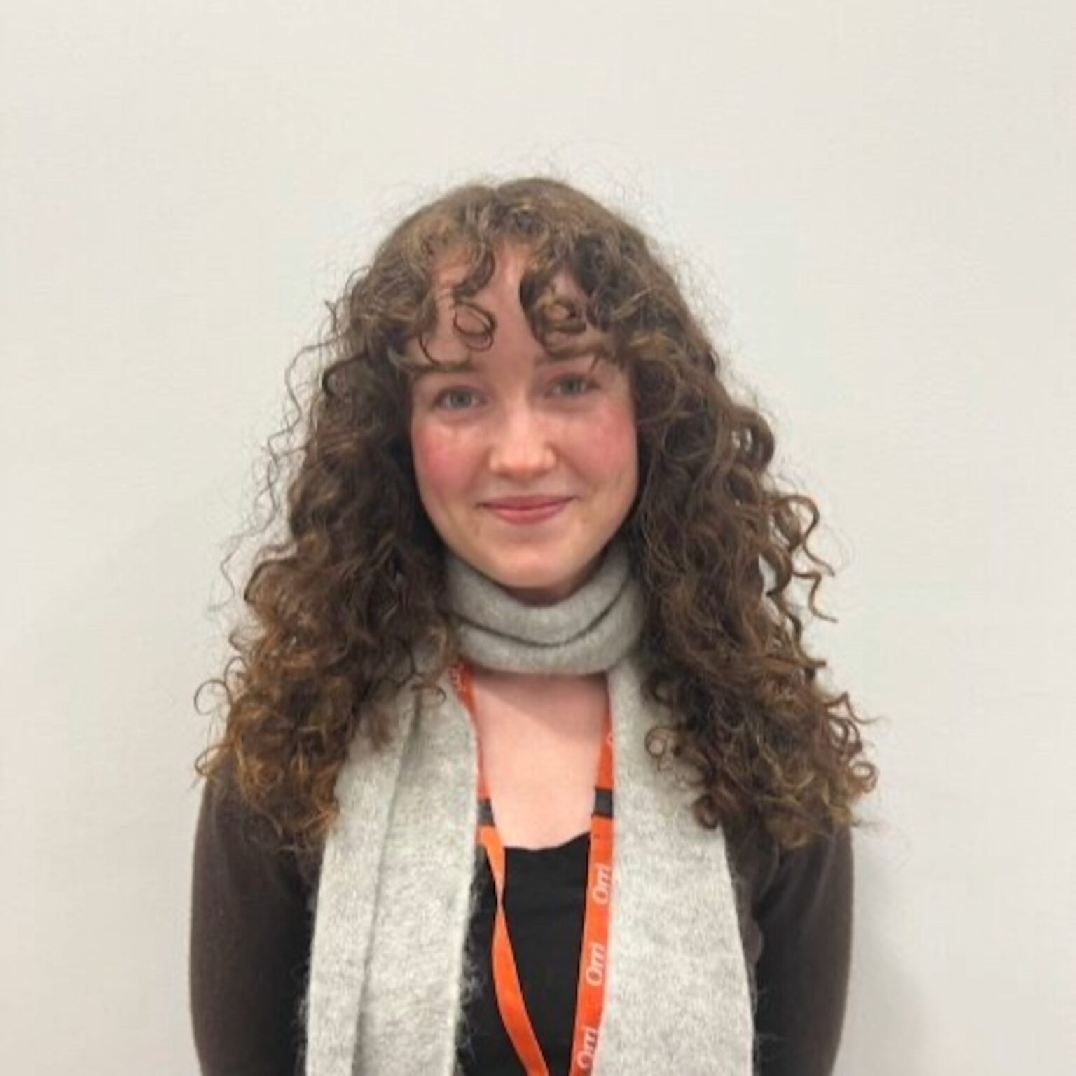 Front-facing portrait of a woman with long curly brown hair, wearing a gray scarf and an orange lanyard, smiling at the camera.