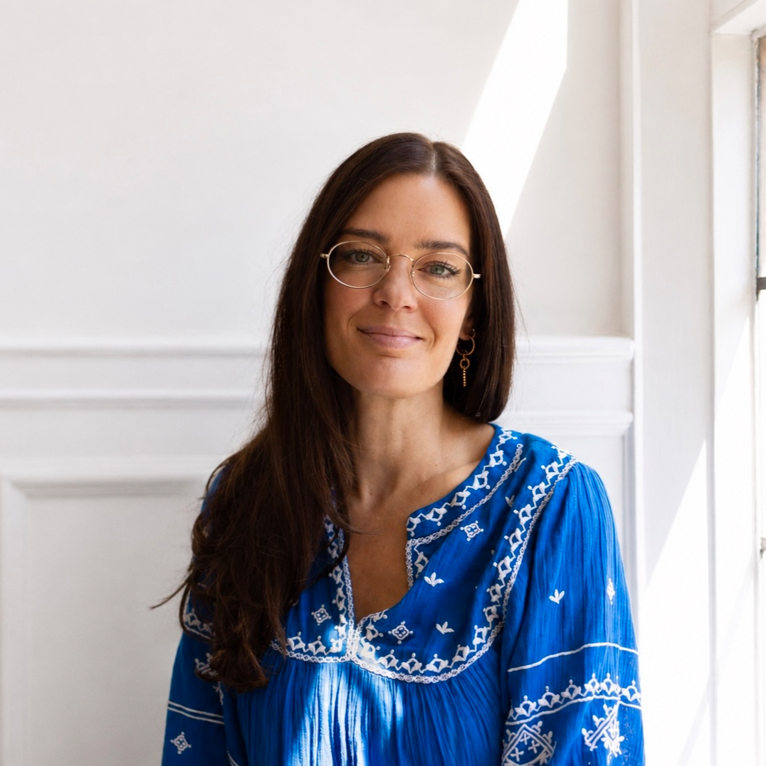 Woman with long brown hair and round glasses, wearing a blue embroidered blouse, smiling by a sunlit window.