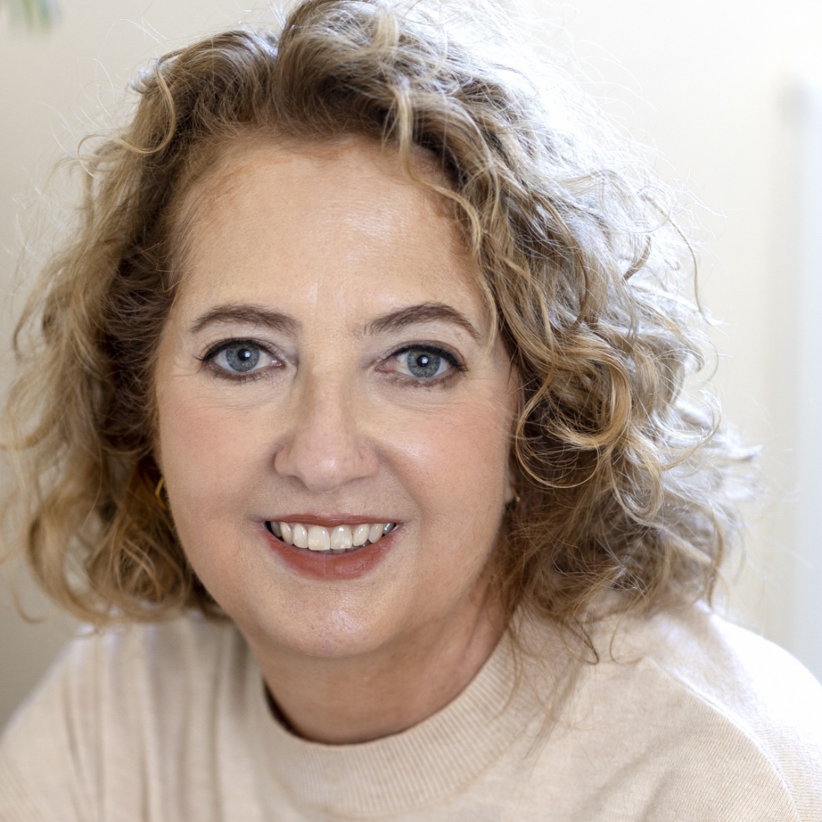 Close-up portrait of a smiling woman with curly light brown hair and blue eyes at the camera angle, wearing a beige top.