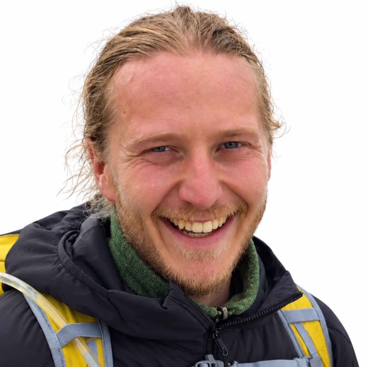 Smiling man with blond hair and stubble wearing a dark jacket and a yellow backpack in an outdoor setting.